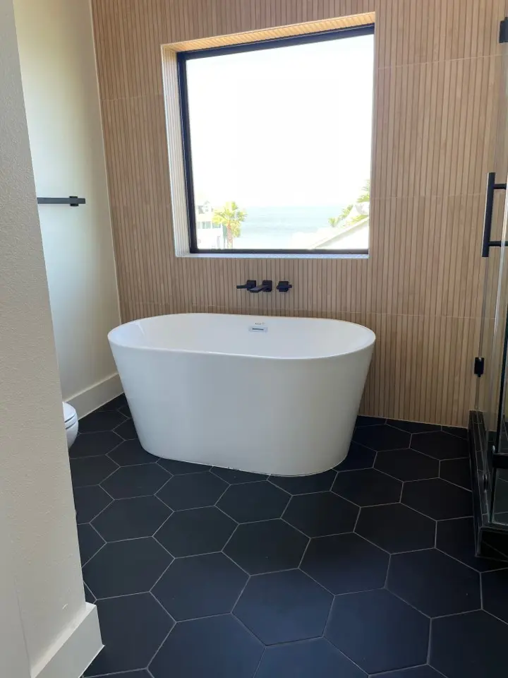 Freestanding soaking tub under a window with wood-look tile accent wall and hexagon floor tile